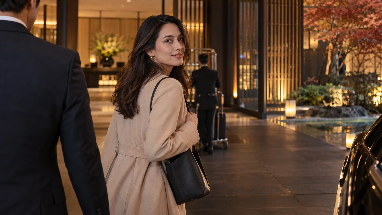 A woman with a shoulder bag looks back while walking near a hotel entrance lined with tall amber-lit columns; a dark handbag is visible beside her. The woman wears a soft cream-colored blouse.