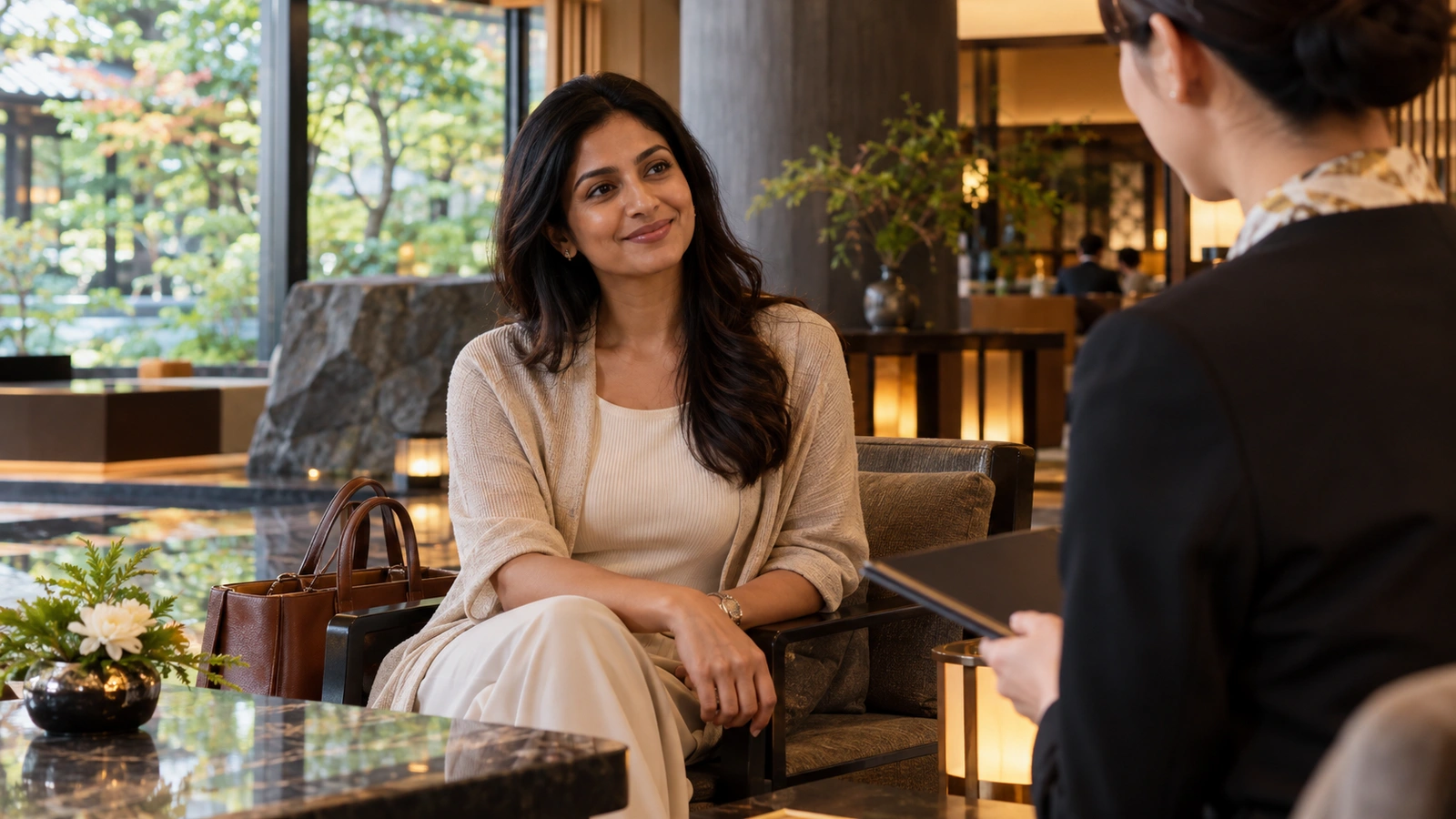 Una mujer habla cara a cara con personal de recepción del hotel en un tranquilo salón de hotel con asientos cómodos y vistas al jardín; junto a ella hay una maleta con ruedas.
