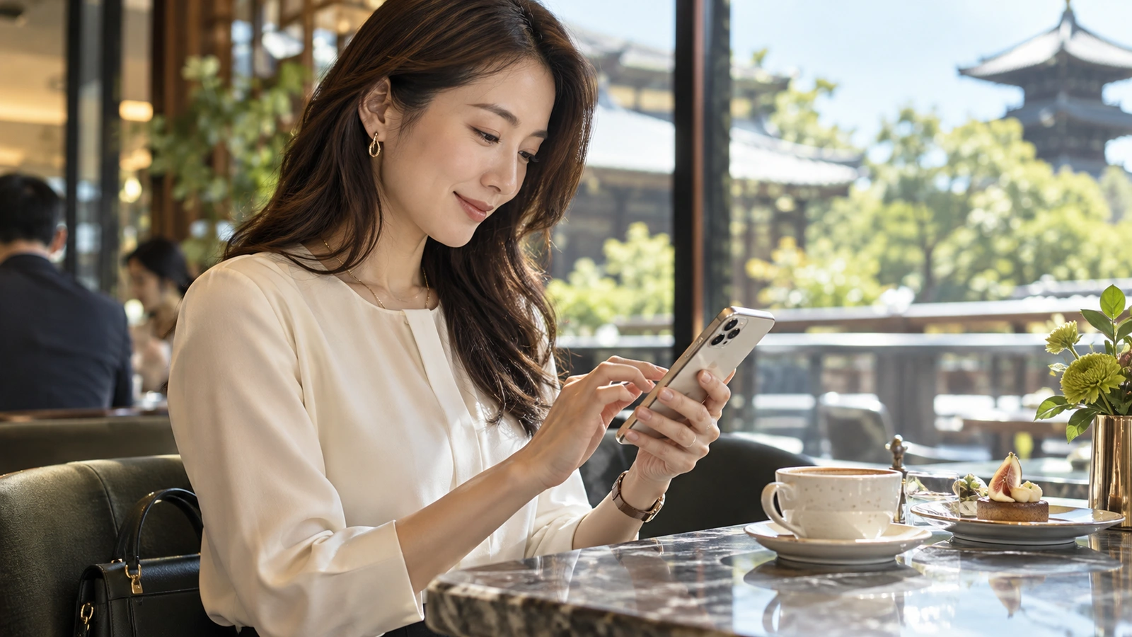 A woman looks down and uses her smartphone inside a bright Japanese café looking out onto traditional roofs and trees; a cup and saucer sit within reach.