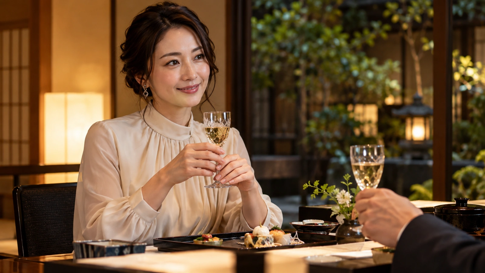 A seated woman smiles while holding a sparkling-wine glass in a warmly lit guest suite beside a landscaped Japanese garden; a tall glass and small flowers are arranged on the table.