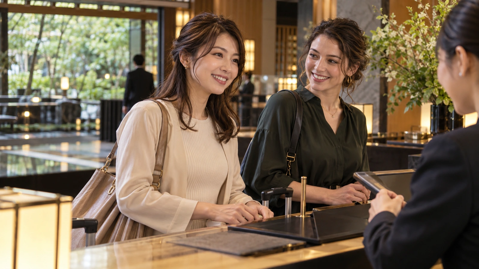 Two women talk with a hotel receptionist across a counter at a polished hotel reception desk beside a landscaped Japanese courtyard; fresh blossoms and table lighting fill the background.