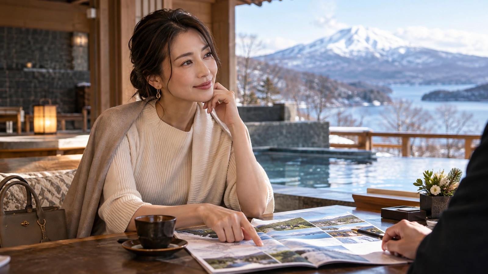 A woman reviews an illustrated brochure with another person in a mountain resort with snowy peaks and a winter lake visible outside; an open photograph-filled brochure lies on the table.