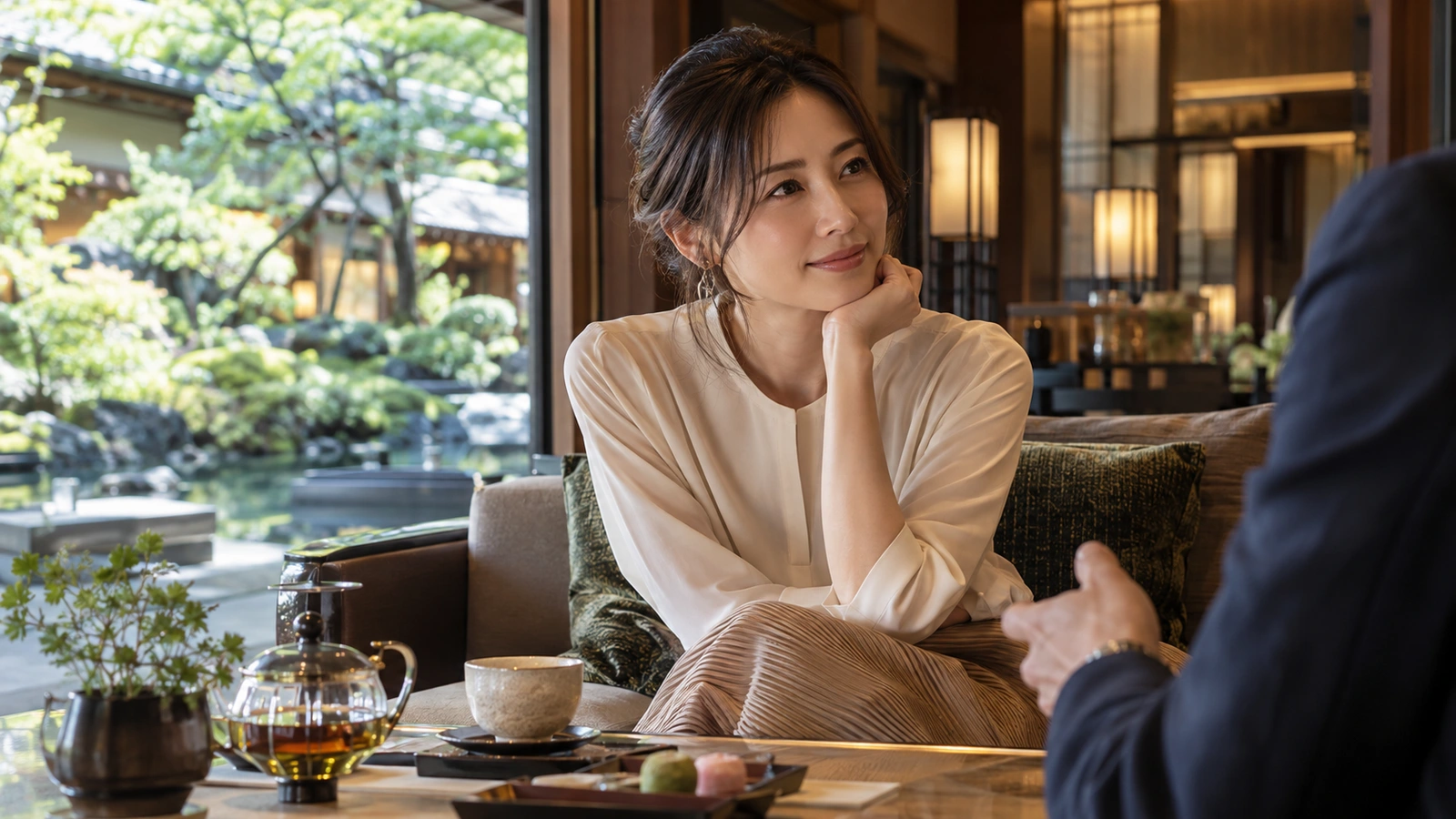 A woman rests her cheek on one hand and listens to a person nearby in a quiet hotel lounge with soft seating and garden greenery; a ceramic cup or teapot completes the quiet table setting.