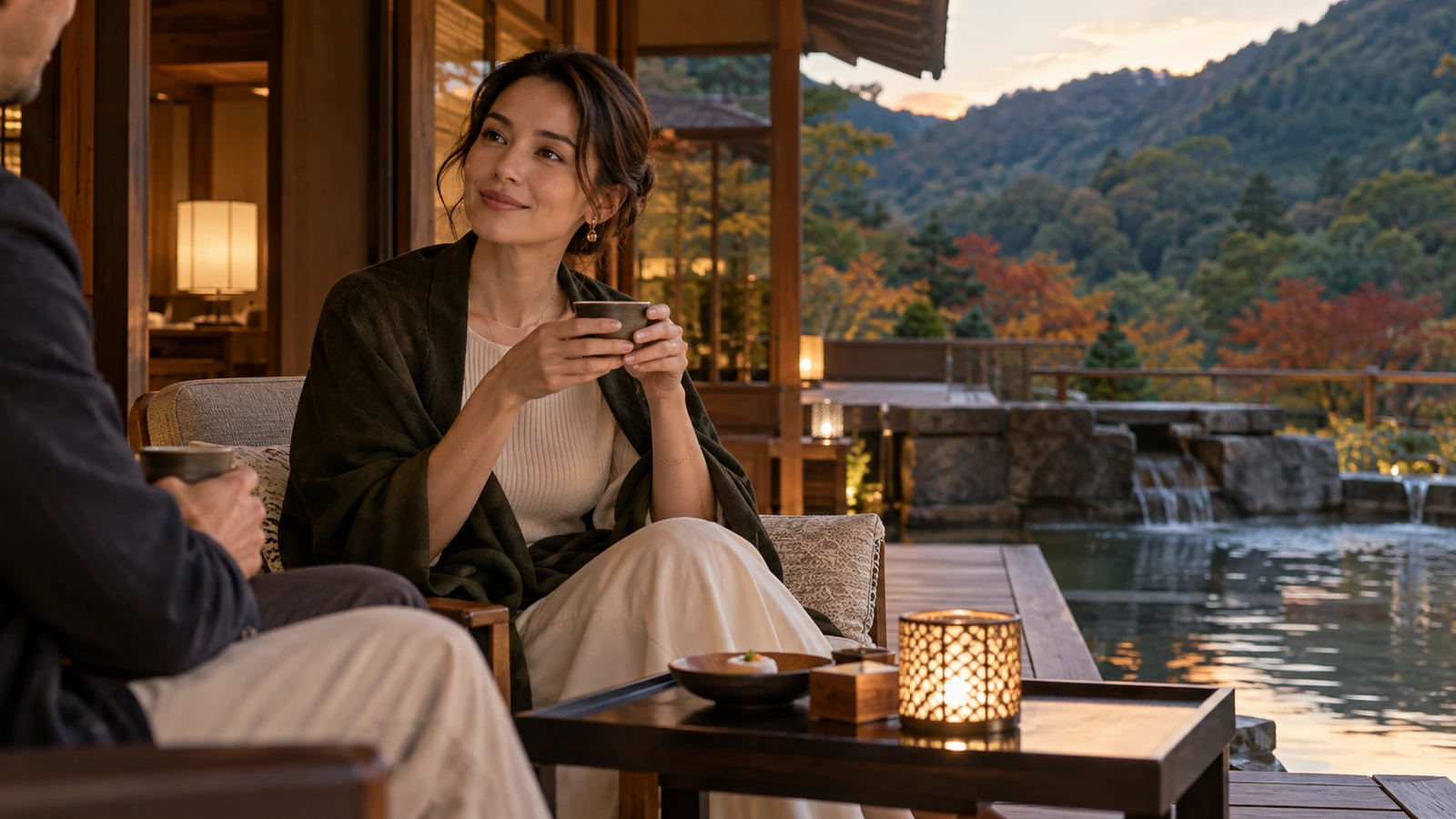 A seated woman holds a ceramic teacup and looks toward the view by an open-air bath with wooded mountains beyond; a ceramic cup or teapot completes the quiet table setting.